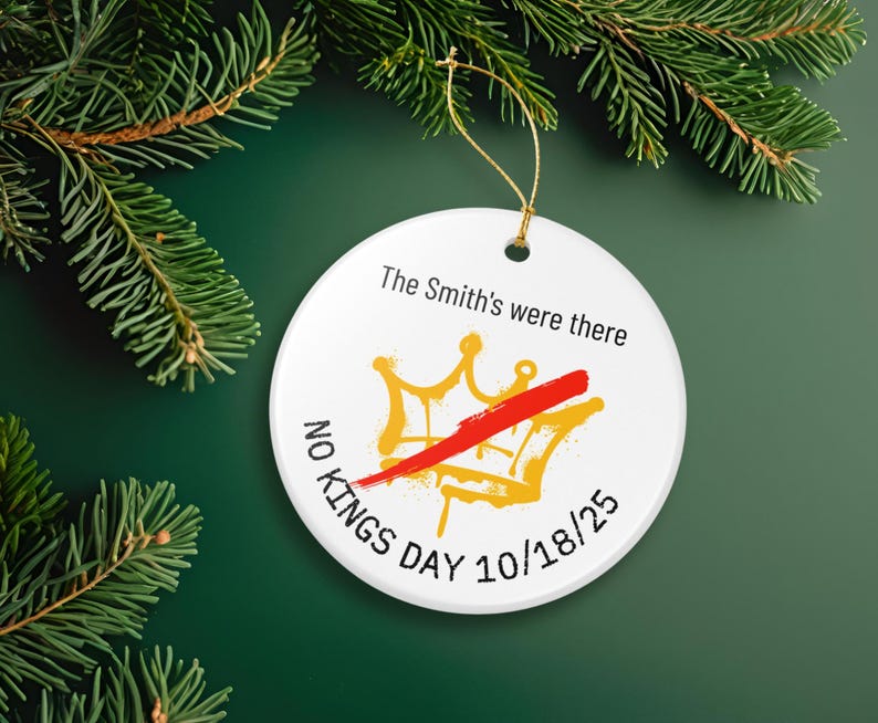 Puede incluir: Adorno navide&ntilde;o redondo de cer&aacute;mica blanca con una cuerda dorada, con un dise&ntilde;o de corona dorada tachada con una raya roja. El texto dice "The Smith's were there", "NO KINGS DAY 10/18/25". Sobre un fondo verde con ramas de pino.