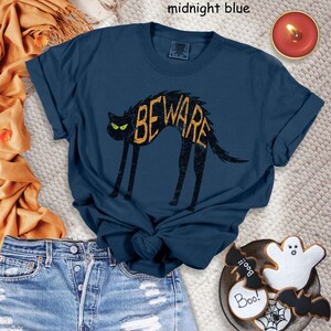 May include: Midnight blue t-shirt featuring a black cat with the word "BEWARE" in a distressed yellow font. The cat has green eyes. The shirt is paired with denim shorts and Halloween-themed cookies.