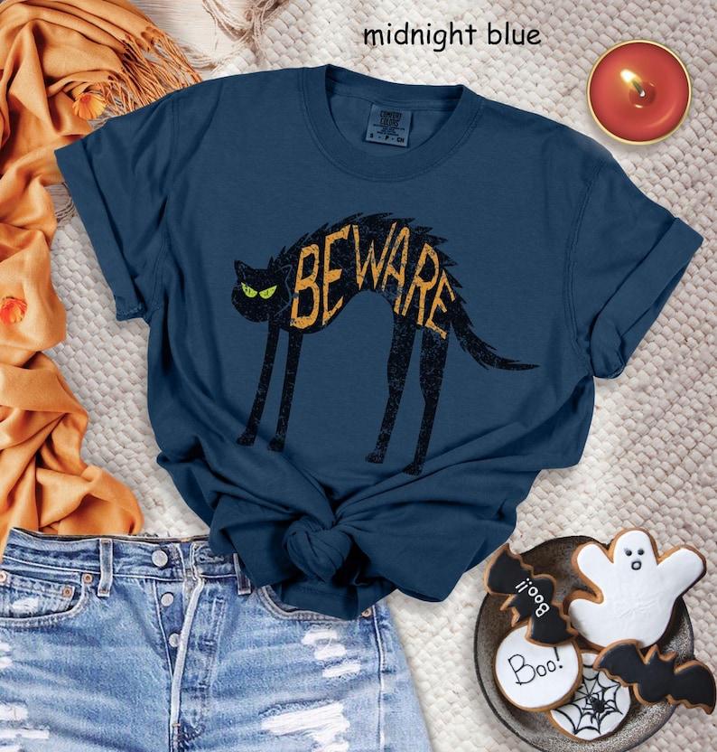 May include: Midnight blue t-shirt featuring a black cat with the word "BEWARE" in a distressed yellow font. The cat has green eyes. The shirt is paired with denim shorts and Halloween-themed cookies.