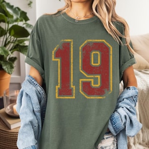 Puede incluir: Camiseta verde oliva con el n&uacute;mero 19 en un dise&ntilde;o desgastado de estilo universitario. El n&uacute;mero es rojo con un contorno amarillo. La camiseta tiene mangas cortas y un corte holgado.