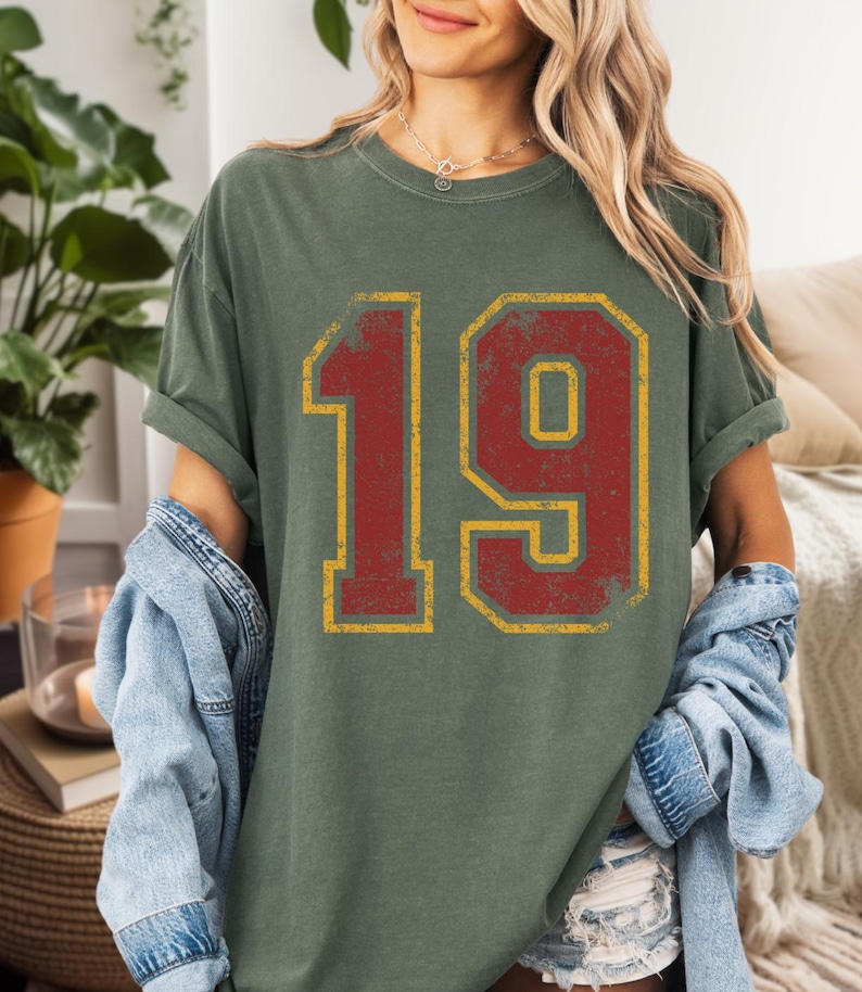 Puede incluir: Camiseta verde oliva con el n&uacute;mero 19 en un dise&ntilde;o desgastado de estilo universitario. El n&uacute;mero es rojo con un contorno amarillo. La camiseta tiene mangas cortas y un corte holgado.