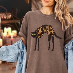 May include: A brown t-shirt with a distressed black cat graphic. The cat has yellow eyes and the word "BEWARE" in yellow letters across its back. The shirt has rolled-up sleeves and a relaxed fit.