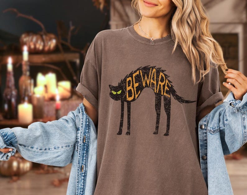 May include: A brown t-shirt with a distressed black cat graphic. The cat has yellow eyes and the word "BEWARE" in yellow letters across its back. The shirt has rolled-up sleeves and a relaxed fit.