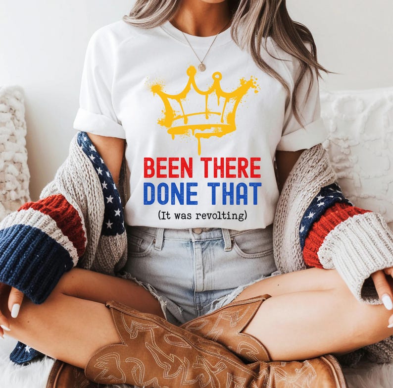 May include: White t-shirt with a yellow crown graphic and the text "BEEN THERE DONE THAT (It was revolting)". The shirt is paired with denim shorts and a patriotic-themed sweater. Brown cowboy boots complete the look.