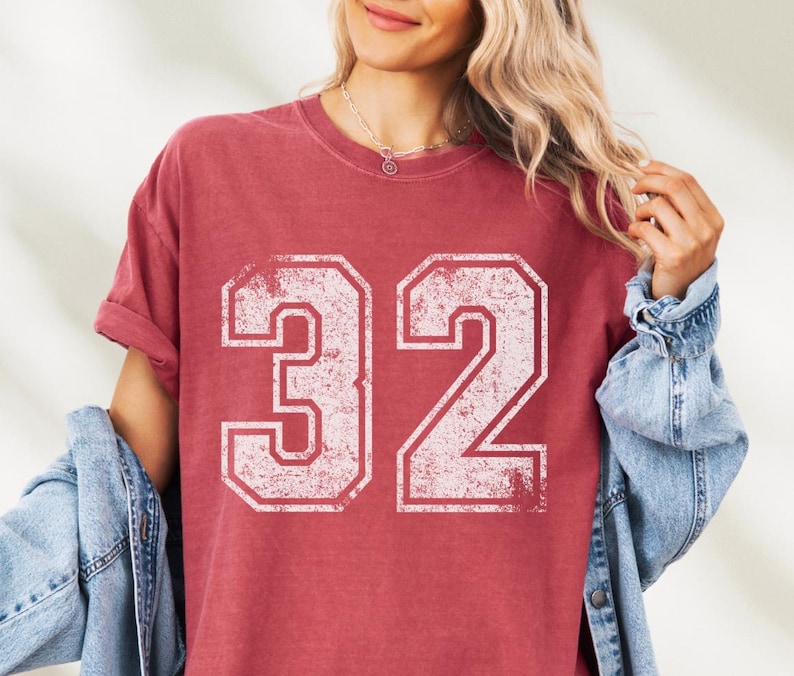 Puede incluir: Camiseta roja con el n&uacute;mero 32 en fuente blanca desgastada. La camiseta tiene mangas cortas y un corte holgado. Se lleva una chaqueta vaquera.