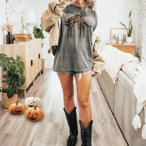 May include: A person wearing a gray t-shirt with a dog graphic and the word "BEWARE". The person is wearing a tan jacket, denim shorts, a hat, and black cowboy boots. Halloween pumpkins are on the floor.