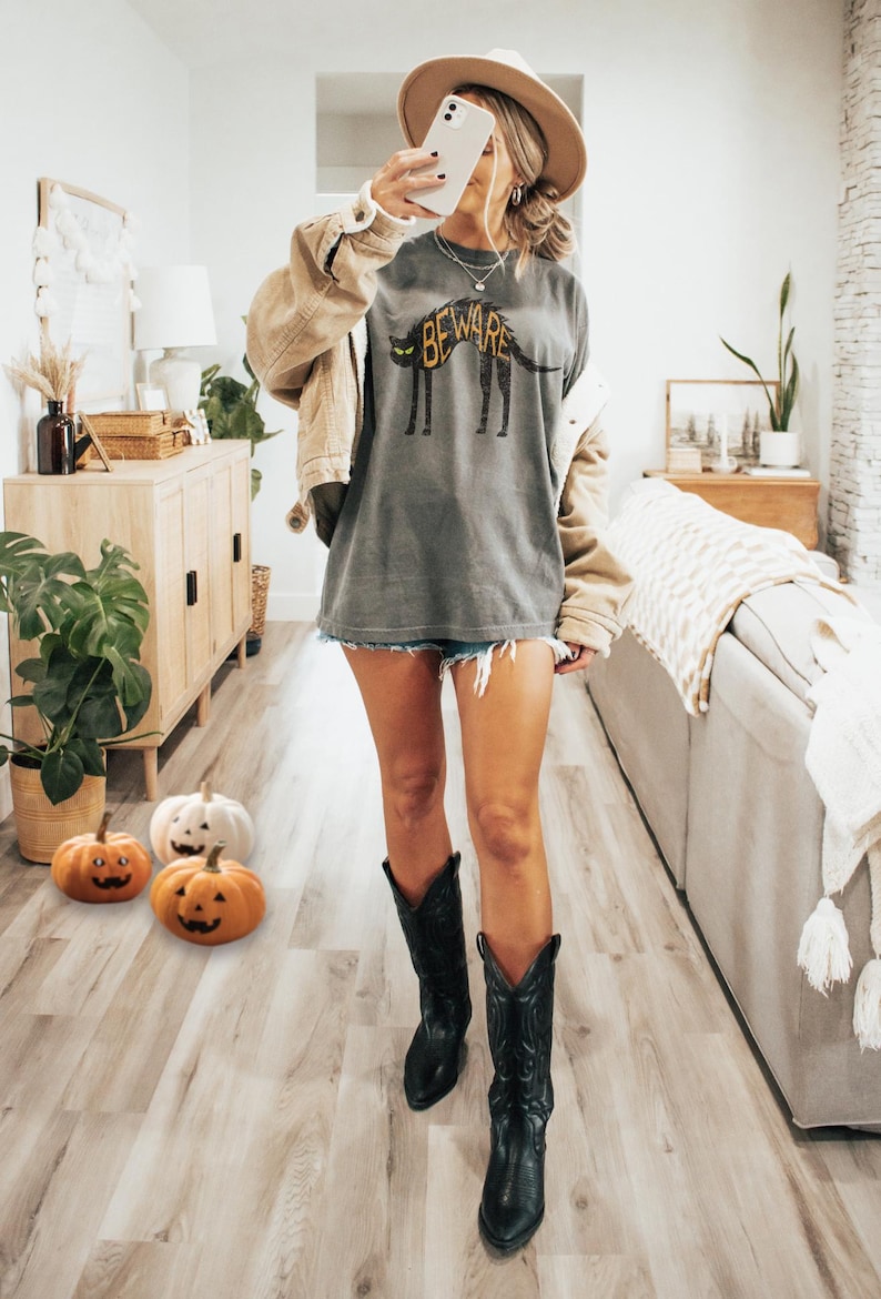 May include: A person wearing a gray t-shirt with a dog graphic and the word "BEWARE". The person is wearing a tan jacket, denim shorts, a hat, and black cowboy boots. Halloween pumpkins are on the floor.