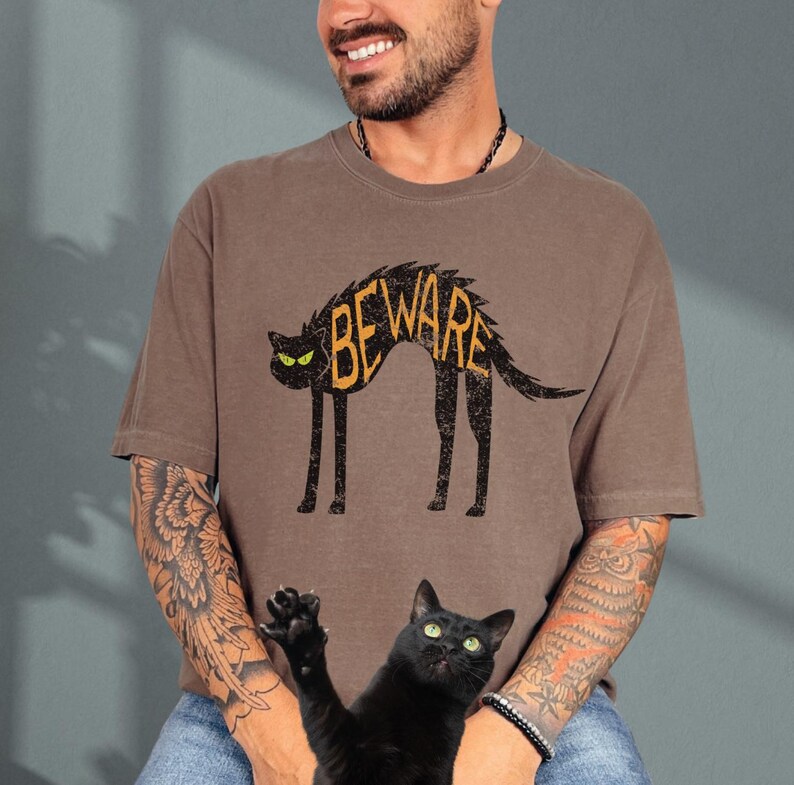 May include: Brown t-shirt with a black cat illustration and the word "BEWARE" in orange. The cat has green eyes and a spiky back. A black cat with green eyes is in the foreground.