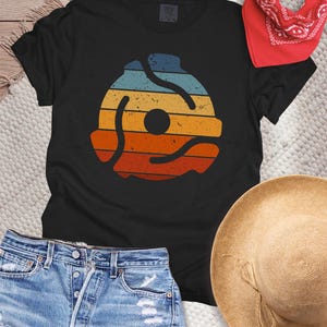May include: A black short-sleeve t-shirt featuring a centered graphic of a 45 RPM record adapter. The design displays horizontal stripes in muted shades of blue, yellow, orange, and red with a distressed, weathered texture throughout the print.