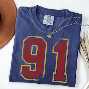 Puede incluir: Una camiseta azul marino con el n&uacute;mero "91" en un dise&ntilde;o rojo y amarillo desgastado. La camiseta tiene cuello redondo y una peque&ntilde;a etiqueta en la parte superior izquierda. Un collar dorado con un colgante descansa sobre la camiseta. Un sombrero marr&oacute;n y una planta seca est&aacute;n en el fondo.
