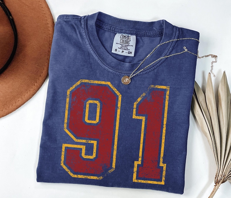 Puede incluir: Una camiseta azul marino con el n&uacute;mero "91" en un dise&ntilde;o rojo y amarillo desgastado. La camiseta tiene cuello redondo y una peque&ntilde;a etiqueta en la parte superior izquierda. Un collar dorado con un colgante descansa sobre la camiseta. Un sombrero marr&oacute;n y una planta seca est&aacute;n en el fondo.