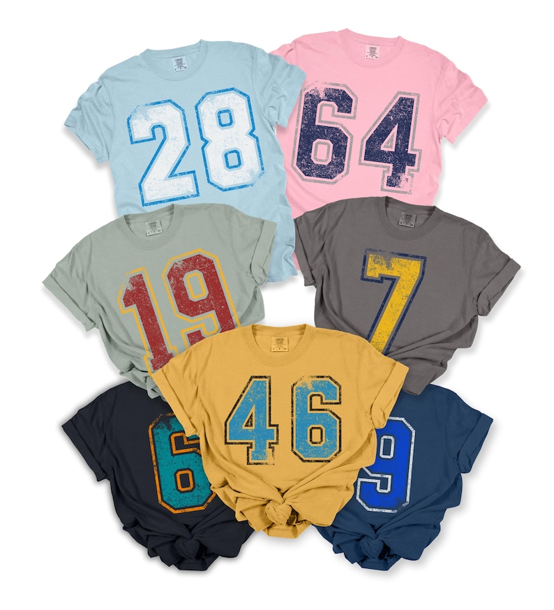 Puede incluir: Una colecci&oacute;n de camisetas de manga corta en varios colores, cada una con un n&uacute;mero grande y desgastado. Los colores incluyen azul claro, rosa, gris, amarillo y azul marino. Los n&uacute;meros son de colores contrastantes, como blanco, azul, rojo y amarillo.