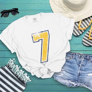 Puede incluir: Camiseta blanca con un gran n&uacute;mero 7 amarillo desgastado, delineado en azul marino. La imagen incluye pantalones cortos de mezclilla, gafas de sol, un sombrero de paja y chanclas a rayas, evocando el verano.