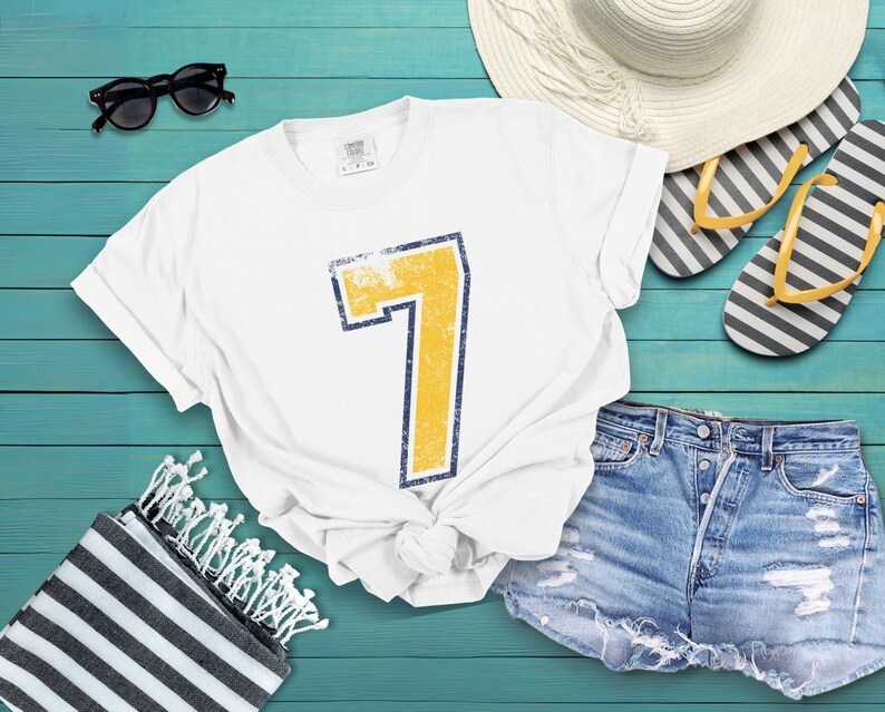 Puede incluir: Camiseta blanca con un gran n&uacute;mero 7 amarillo desgastado, delineado en azul marino. La imagen incluye pantalones cortos de mezclilla, gafas de sol, un sombrero de paja y chanclas a rayas, evocando el verano.