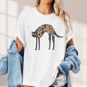 May include: White t-shirt featuring a distressed black cat graphic with the word "BEWARE" in orange. The cat has green eyes and a curved back. The shirt has rolled-up sleeves and a casual fit.
