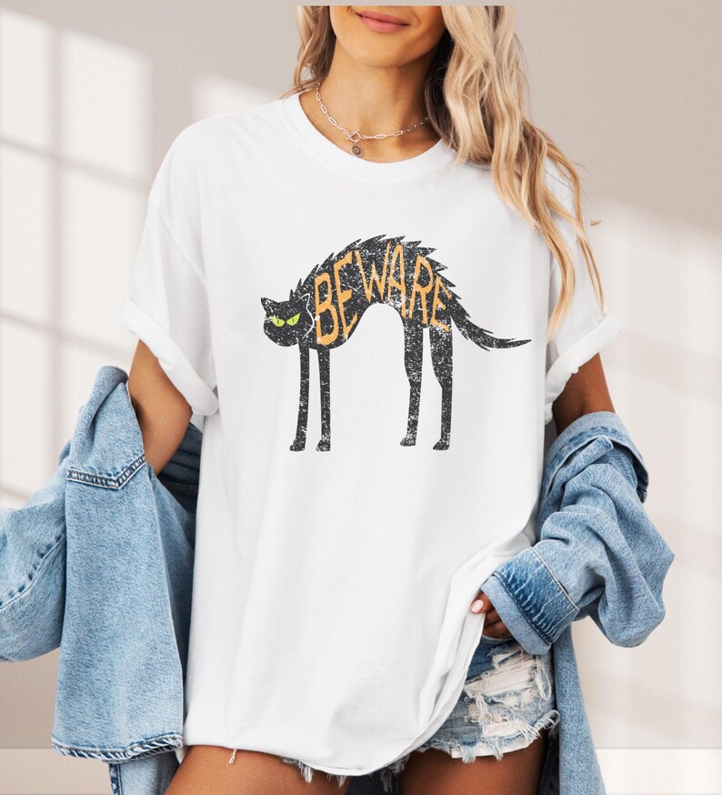 May include: White t-shirt featuring a distressed black cat graphic with the word "BEWARE" in orange. The cat has green eyes and a curved back. The shirt has rolled-up sleeves and a casual fit.