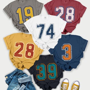 Puede incluir: Una colecci&oacute;n de camisetas de estilo vintage en varios colores, cada una con un n&uacute;mero grande. Las camisetas son grises, rojas, blancas, doradas, azul marino y azul oscuro. Los n&uacute;meros tienen un aspecto envejecido. Tambi&eacute;n hay vaqueros y zapatillas amarillas.