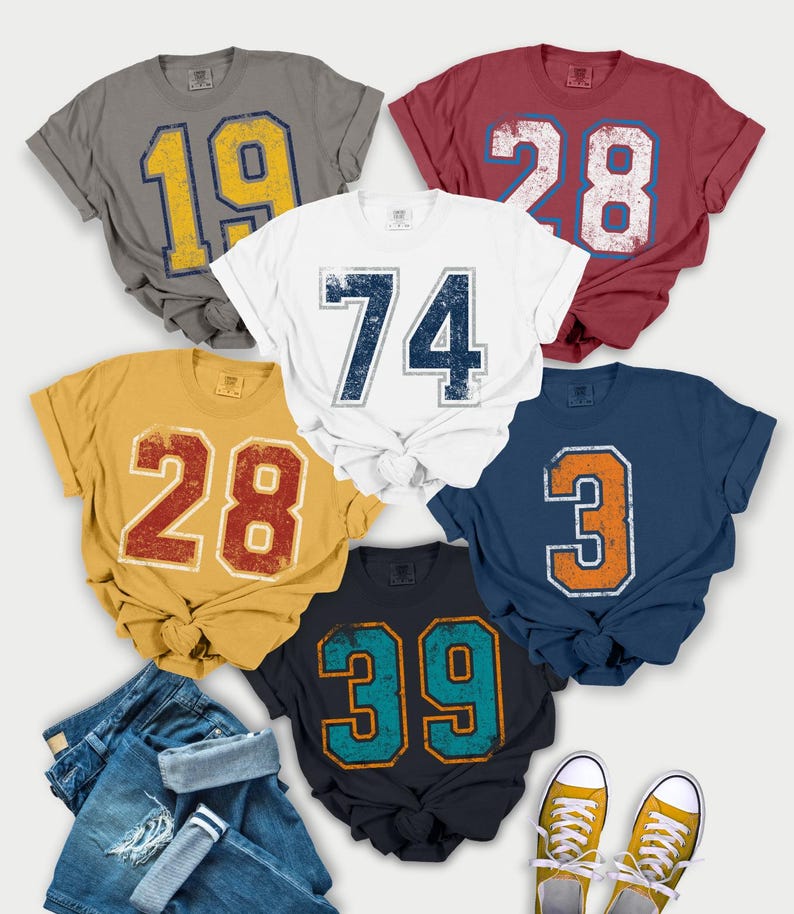 Puede incluir: Una colecci&oacute;n de camisetas de estilo vintage en varios colores, cada una con un n&uacute;mero grande. Las camisetas son grises, rojas, blancas, doradas, azul marino y azul oscuro. Los n&uacute;meros tienen un aspecto envejecido. Tambi&eacute;n hay vaqueros y zapatillas amarillas.