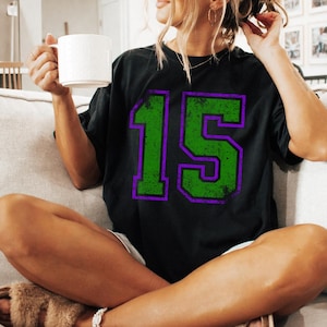 Puede incluir: Camiseta negra con un gran n&uacute;mero 15 verde desgastado con contorno morado. La persona que lleva la camiseta est&aacute; sentada en un sof&aacute; y sostiene una taza blanca. El estilo es informal y relajado.