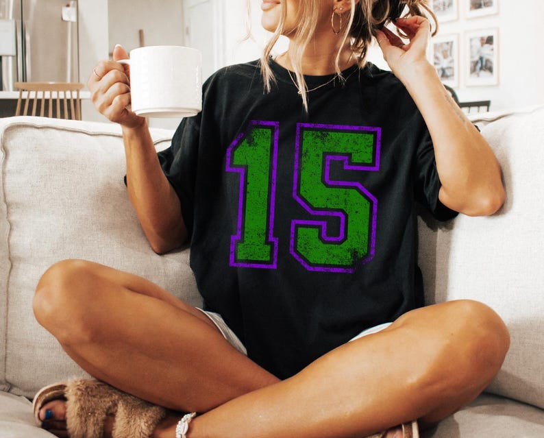 Puede incluir: Camiseta negra con un gran n&uacute;mero 15 verde desgastado con contorno morado. La persona que lleva la camiseta est&aacute; sentada en un sof&aacute; y sostiene una taza blanca. El estilo es informal y relajado.