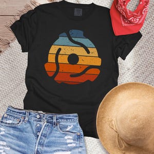 May include: Black t-shirt featuring a retro-style graphic of a 45 RPM record adapter in shades of blue, yellow, orange, and red. The shirt is paired with denim shorts, a red bandana, and a straw hat, creating a casual, summery aesthetic.
