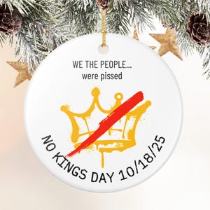 Puede incluir: Adorno navide&ntilde;o redondo de cer&aacute;mica blanca con cord&oacute;n dorado. El adorno presenta el texto "WE THE PEOPLE... were pissed" sobre una corona dorada tachada con una raya roja. Las palabras "NO KINGS DAY 10/18/25" est&aacute;n impresas en la parte inferior.
