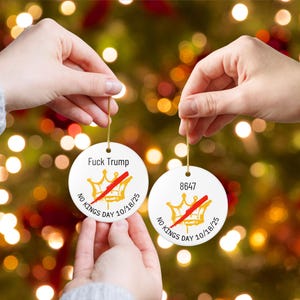 Puede incluir: Dos adornos navide&ntilde;os redondos de cer&aacute;mica blanca con hilo dorado. Cada uno presenta un dise&ntilde;o de corona dorada tachada en rojo, con los textos "Fuck Trump", "8647" y "NO KINGS DAY 10/18/25". Los adornos se sostienen contra un fondo borroso de luces navide&ntilde;as.