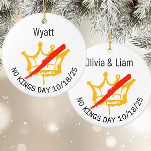 Puede incluir: Dos adornos navide&ntilde;os redondos de cer&aacute;mica blanca con hilo dorado. Cada adorno presenta un dise&ntilde;o de corona dorada tachado con una barra roja, el texto "NO KINGS DAY 10/18/25" y un nombre. Uno dice "Wyatt", el otro "Olivia & Liam".