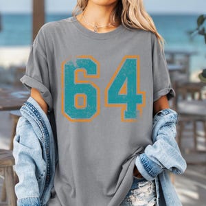 Puede incluir: Camiseta gris con el n&uacute;mero "64" en turquesa con contorno naranja, con un aspecto desgastado. La modelo lleva una chaqueta vaquera y unos shorts.