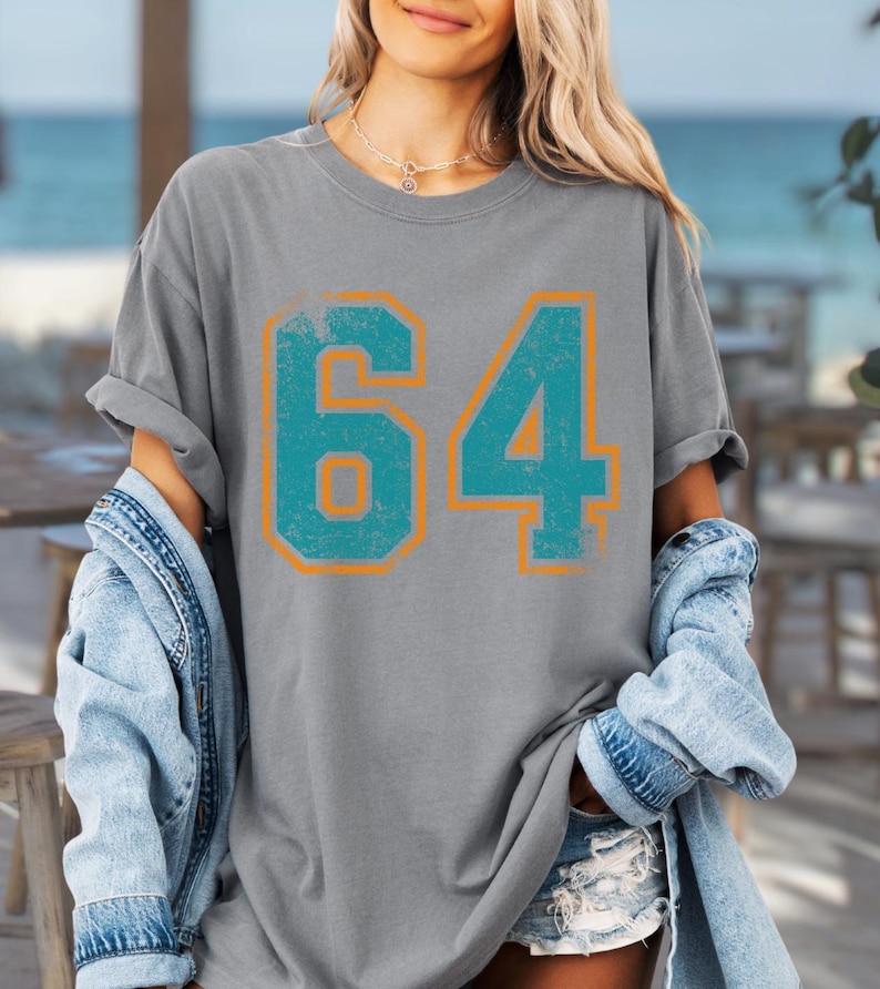 Puede incluir: Camiseta gris con el n&uacute;mero "64" en turquesa con contorno naranja, con un aspecto desgastado. La modelo lleva una chaqueta vaquera y unos shorts.