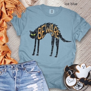 May include: Ice blue t-shirt featuring a black cat with the word "BEWARE" in orange. The cat has green eyes and a distressed design. The shirt is paired with denim shorts and Halloween-themed cookies.