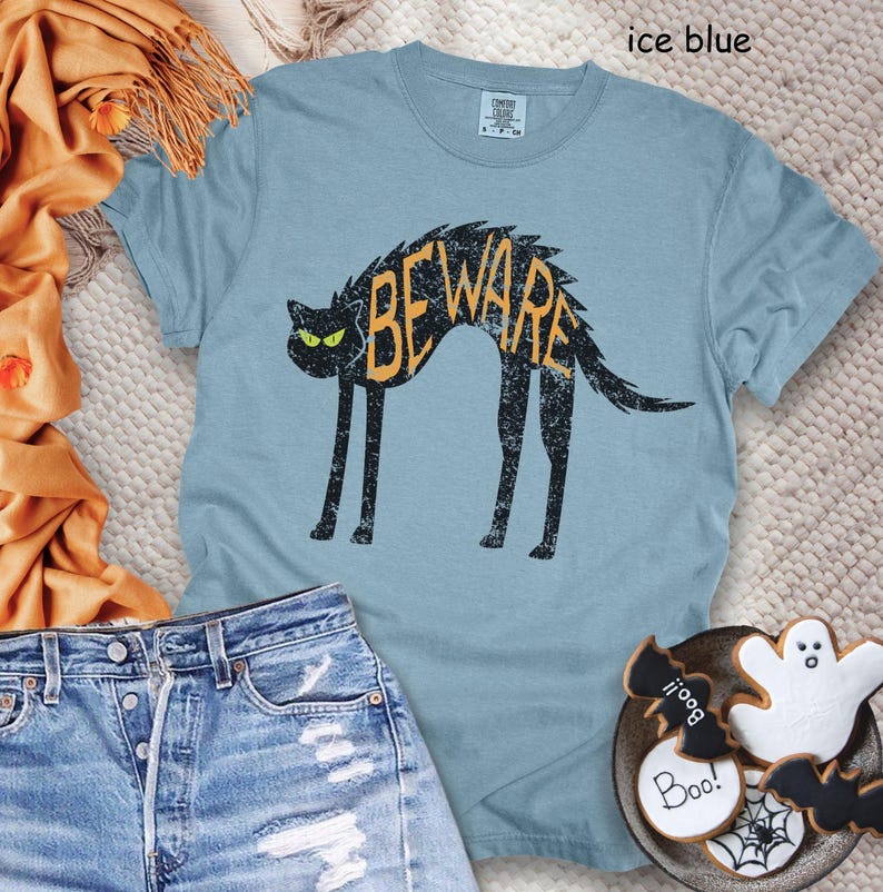 May include: Ice blue t-shirt featuring a black cat with the word "BEWARE" in orange. The cat has green eyes and a distressed design. The shirt is paired with denim shorts and Halloween-themed cookies.