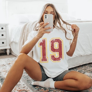 May include: A white short-sleeved t-shirt featuring the number 19 printed in a distressed red font with a yellow outline. The shirt is paired with dark grey athletic shorts and white socks, creating a casual look suitable for everyday wear.