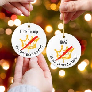 Puede incluir: Dos adornos navide&ntilde;os redondos de cer&aacute;mica blanca con hilo dorado. Uno tiene el texto "Fuck Trump" sobre una corona dorada con una raya roja y el texto "NO KINGS DAY 10/18/25". El otro tiene el n&uacute;mero "8647" y una corona similar.