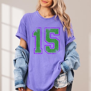 Puede incluir: Camiseta lila con el n&uacute;mero "15" impreso en verde con contorno morado. El n&uacute;mero tiene un aspecto desgastado. La camiseta de manga corta tiene un corte holgado, ideal para un look casual.