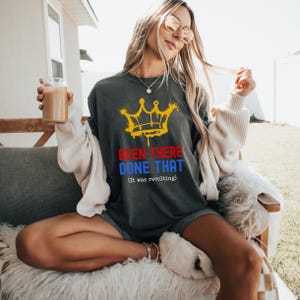 May include: Dark gray t-shirt with a yellow crown graphic and the text "BEEN THERE DONE THAT (it was revolting)" in red and blue. The shirt is worn by a person sitting outdoors.