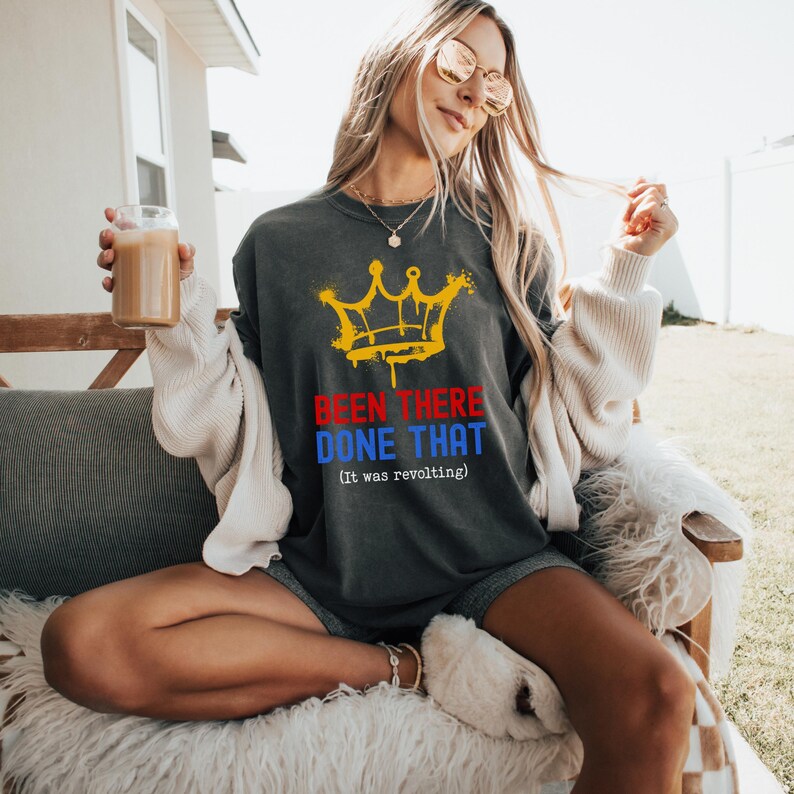 May include: Dark gray t-shirt with a yellow crown graphic and the text "BEEN THERE DONE THAT (it was revolting)" in red and blue. The shirt is worn by a person sitting outdoors.