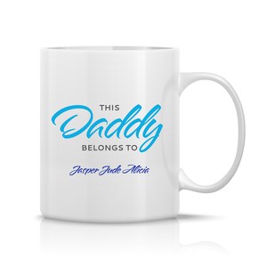 Pode incluir: Caneca de cerâmica branca com uma alça curva. A caneca apresenta o texto "THIS Daddy BELONGS TO" em azul, seguido pelos nomes "Jasper Jude Alicia" em uma fonte azul menor. A caneca é projetada para presentear.