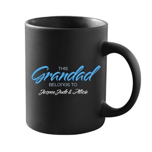 Pode incluir: Caneca de cerâmica preta com uma alça curva. A caneca apresenta o texto "This Grandad belongs to Jasper Jude & Alicia" em escrita azul e branca. A caneca foi projetada para presentear.