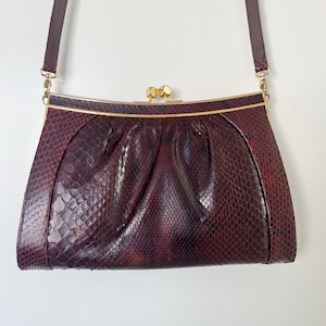 May include: A vintage burgundy handbag with a snakeskin pattern. The bag has a gold-toned clasp and trim, and a matching shoulder strap. The design includes a pleated front and a structured shape, showcasing a classic, elegant style.