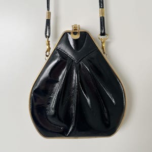 May include: A black patent leather handbag with a structured, trapezoidal shape and a gold-tone metal frame. The bag features a central clasp closure, pleated detailing on the front surface, and a thin, matching black shoulder strap with gold accents.