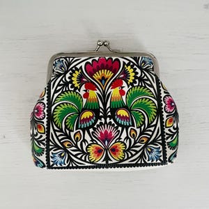 Könnte beinhalten: Eine weiße Geldbörse mit Metallverschluss und farbenfrohem Volkskunstdesign. Das Design umfasst zwei Hähne, Blumen und Blätter in Rot, Grün, Gelb und Blau. Die Geldbörse hat eine rechteckige Form mit abgerundeten Kanten.