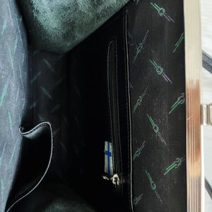 May include: The inside of a black leather purse with a silver clasp. The lining is black with a repeating pattern of green and white lines. There is a zipped pocket on the right side of the image.