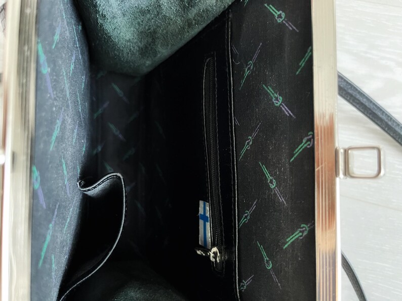 May include: The inside of a black leather purse with a silver clasp. The lining is black with a repeating pattern of green and white lines. There is a zipped pocket on the right side of the image.