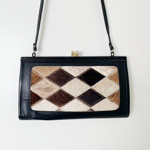 May include: A rectangular black handbag featuring a central panel with a diamond-patterned patchwork of brown, tan, and white hair-on hide. The bag has a thin black shoulder strap, a black frame, and a small metallic clasp closure at the top centre.