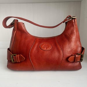 May include: A reddish-brown leather shoulder bag with a curved shape and a single handle. The bag has a textured surface, buckle closures on each side, and a small embossed logo on the front.