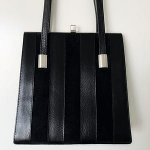 May include: A black leather handbag with a rectangular shape and vertical stripes. The bag has two silver clasps on the sides and a silver clasp on the top.