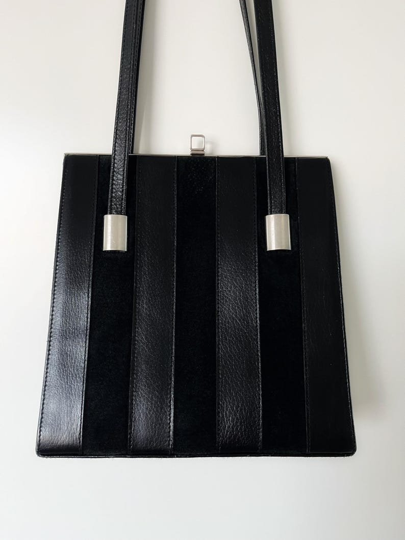 May include: A black leather handbag with a rectangular shape and vertical stripes. The bag has two silver clasps on the sides and a silver clasp on the top.