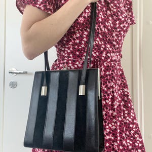 May include: A black leather handbag with a silver clasp and vertical stripes. The bag has a long strap and is being held by a person wearing a floral dress.