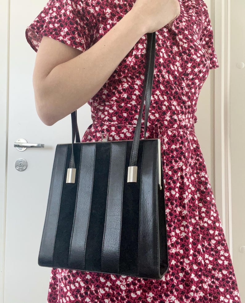 May include: A black leather handbag with a silver clasp and vertical stripes. The bag has a long strap and is being held by a person wearing a floral dress.